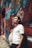 A person with headphones around their neck leans against a graffiti-covered brick wall in an alley. They are holding a white bag and wearing a white T-shirt. The scene is brightly lit with colorful urban art surrounding them.
