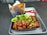A serving of fried chicken pieces drizzled with a glossy sauce and garnished with chopped green onions and broccoli florets is placed in a black container. This is accompanied by a neatly packaged spring roll placed in a branded paper holder. The ensemble is set on a silver tray with a red plastic spoon and a white napkin.