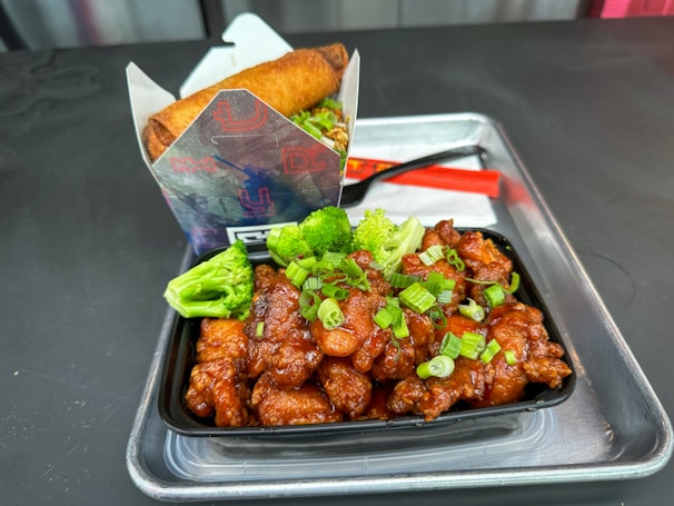 A serving of fried chicken pieces drizzled with a glossy sauce and garnished with chopped green onions and broccoli florets is placed in a black container. This is accompanied by a neatly packaged spring roll placed in a branded paper holder. The ensemble is set on a silver tray with a red plastic spoon and a white napkin.