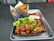 A serving of fried chicken pieces drizzled with a glossy sauce and garnished with chopped green onions and broccoli florets is placed in a black container. This is accompanied by a neatly packaged spring roll placed in a branded paper holder. The ensemble is set on a silver tray with a red plastic spoon and a white napkin.
