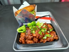 A serving of fried chicken pieces drizzled with a glossy sauce and garnished with chopped green onions and broccoli florets is placed in a black container. This is accompanied by a neatly packaged spring roll placed in a branded paper holder. The ensemble is set on a silver tray with a red plastic spoon and a white napkin.