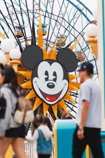 A large Ferris wheel features a prominent face of a cartoon mouse at its center. People in casual attire walk around the foreground, partially obscuring parts of the scene. The wheel's vibrant gondolas are visible, as well as decorative elements like stylized sun rays surrounding the mouse face.