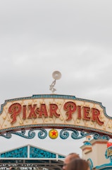 A decorative sign reads 'Pixar Pier' in bold, illuminated letters, with intricate patterns and star motifs. Above the sign, a figure of a lamp is positioned. The sky is overcast with a light grey hue, and parts of a building can be seen on the right side.