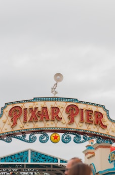 A decorative sign reads 'Pixar Pier' in bold, illuminated letters, with intricate patterns and star motifs. Above the sign, a figure of a lamp is positioned. The sky is overcast with a light grey hue, and parts of a building can be seen on the right side.