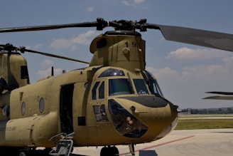 A military helicopter, likely a CH-47 Chinook, is parked on an airfield. It has a large, olive-green body with tandem rotors. The landing gear is visible, and the helicopter appears to have distinctive windows at the cockpit. The background shows a clear sky with some clouds.