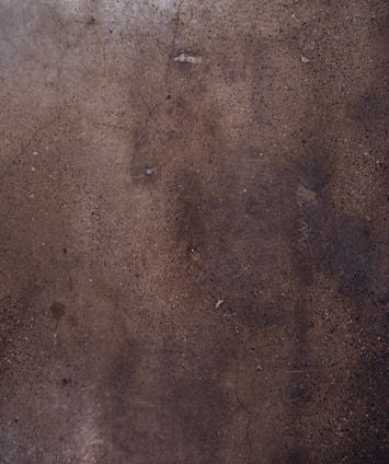 A textured concrete surface with various shades of brown and gray, displaying small specks and some cracks throughout the surface.