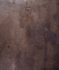 A textured concrete surface with various shades of brown and gray, displaying small specks and some cracks throughout the surface.