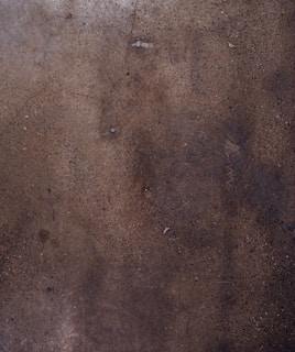 A textured concrete surface with various shades of brown and gray, displaying small specks and some cracks throughout the surface.