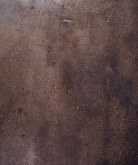 A textured concrete surface with various shades of brown and gray, displaying small specks and some cracks throughout the surface.