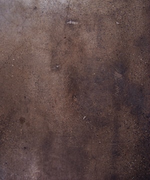 A textured concrete surface with various shades of brown and gray, displaying small specks and some cracks throughout the surface.