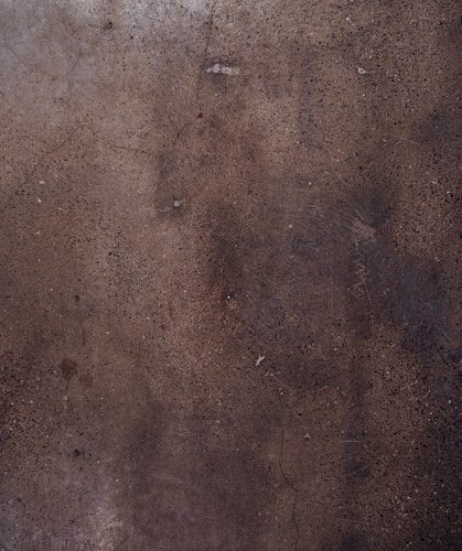 A textured concrete surface with various shades of brown and gray, displaying small specks and some cracks throughout the surface.