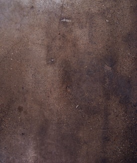 A textured concrete surface with various shades of brown and gray, displaying small specks and some cracks throughout the surface.
