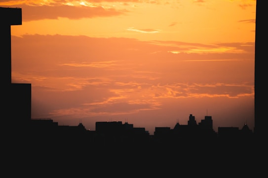 A city skyline silhouetted against a vibrant orange and yellow sunset. The sky is partially covered with some clouds, adding texture to the sunset. There are tall buildings with distinct shapes most noticeable at the left, center, and right edges of the image, creating a contrast between the dark silhouettes and the bright sky.
