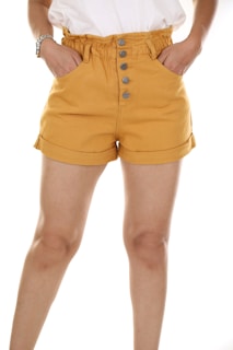 A person is wearing mustard-colored high-waisted shorts with buttons and pockets. The shorts have a rolled-up hem and are paired with a white shirt.