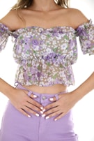 A person wearing a floral off-the-shoulder top with ruffled short sleeves and high-waisted lilac pants. The top showcases a pattern of purple and white flowers. The person's hands are resting on the waistband of the pants, fingernails are painted white.