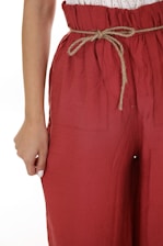 A close-up view of a person wearing loose-fitting red pants with a drawstring waistband. The pants have a crinkled texture, and the drawstring is made of a natural fiber material. The person’s hand is partially visible, resting on the side of the pants.