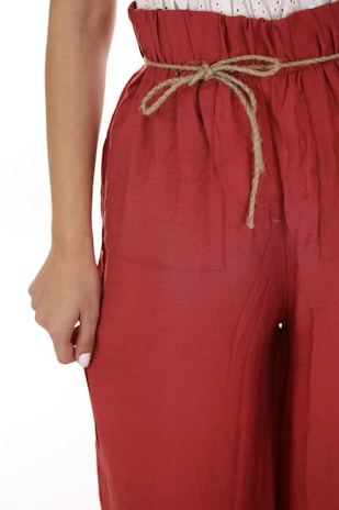 A close-up view of a person wearing loose-fitting red pants with a drawstring waistband. The pants have a crinkled texture, and the drawstring is made of a natural fiber material. The person’s hand is partially visible, resting on the side of the pants.