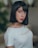 A person with straight, dark hair styled in a bob is wearing a white top with the word 'MANGO' printed on it. The background is softly blurred, suggesting an indoor setting.