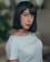 A person with straight, dark hair styled in a bob is wearing a white top with the word 'MANGO' printed on it. The background is softly blurred, suggesting an indoor setting.