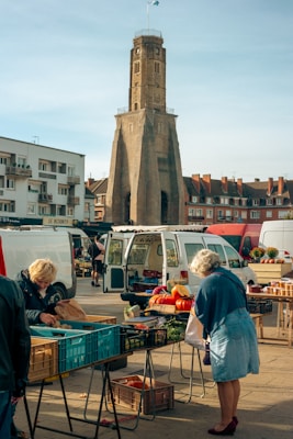A bustling outdoor market with vendors setting up stalls and selling goods. In the foreground, an elderly woman examines items on a table, while in the background are large vans and a variety of products including fruits and vegetables. A historic tower stands prominently, surrounded by residential buildings under a clear sky.