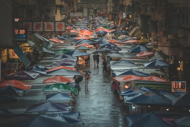 A bustling street market scene with numerous stalls covered by colorful tarps lining each side of a narrow street. People are walking, some with umbrellas, suggesting it is a rainy day. The atmosphere is lively, with bright signs and advertisements in a script providing a cultural context.