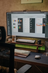 A workspace with a computer monitor displaying design elements labeled as color, typography, and effects. Below the monitor are two mechanical keyboards on a dual-tiered wooden stand, alongside a mouse and a candle. The room has a neutral tone with a touch of green ambient lighting.