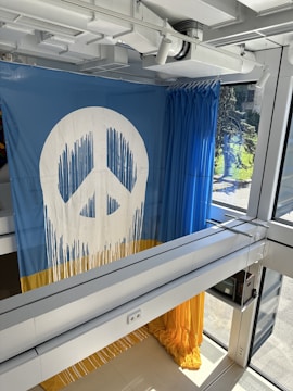 A large mural depicting a peace symbol dominates the scene. The symbol is white and has drips extending downward, resembling paint. The lower half of the mural features a bold yellow stripe, creating a strong contrast with the blue background. This interior view shows part of a modern building with large windows and visible ceiling ducts.
