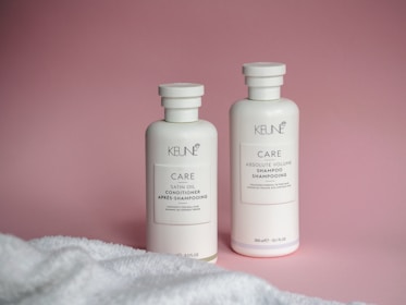 Two white bottles of Keune hair care products, including a conditioner and a shampoo, are placed against a pink backdrop. A white towel is partially visible in the foreground.