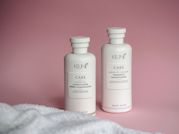 Two white bottles of Keune hair care products, including a conditioner and a shampoo, are placed against a pink backdrop. A white towel is partially visible in the foreground.