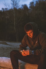 A young person wearing a hoodie is sitting outdoors in a forested area at dusk, focused on a smartphone in their hands. The soft glow from the screen lights up their face as they are seated on a log.