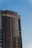 A tall, modern apartment building with a combination of brick and light-colored siding reaches into a clear blue sky. The structure features numerous windows and a few balconies. The design is sleek and contemporary, emphasizing vertical lines.