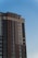 A tall, modern apartment building with a combination of brick and light-colored siding reaches into a clear blue sky. The structure features numerous windows and a few balconies. The design is sleek and contemporary, emphasizing vertical lines.