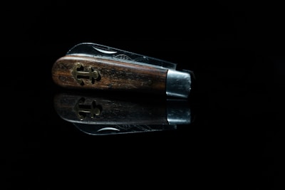 A close-up view of a folded pocket knife with a wooden handle, featuring a decorative cross-shaped metal inlay. The knife is set against a black background, with a reflection visible beneath it.