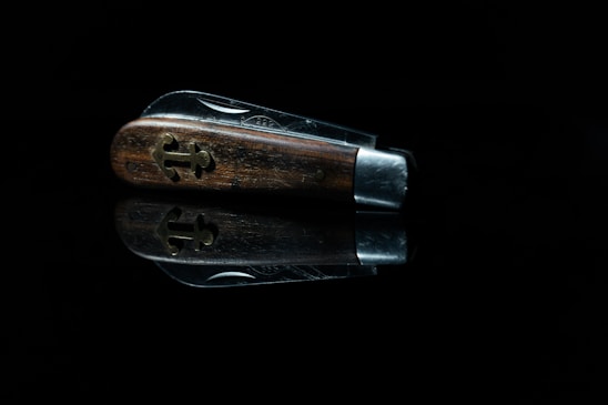 A close-up view of a folded pocket knife with a wooden handle, featuring a decorative cross-shaped metal inlay. The knife is set against a black background, with a reflection visible beneath it.