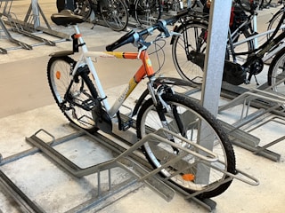 A bicycle is parked in an indoor bike rack among several other bikes. It features a white frame with orange accents and is secured in a metal rack on a concrete floor. The handlebars have black grips, and the tires are designed for outdoor terrain.