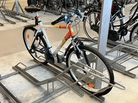 A bicycle is parked in an indoor bike rack among several other bikes. It features a white frame with orange accents and is secured in a metal rack on a concrete floor. The handlebars have black grips, and the tires are designed for outdoor terrain.