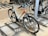 A bicycle is parked in an indoor bike rack among several other bikes. It features a white frame with orange accents and is secured in a metal rack on a concrete floor. The handlebars have black grips, and the tires are designed for outdoor terrain.