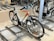 A bicycle is parked in an indoor bike rack among several other bikes. It features a white frame with orange accents and is secured in a metal rack on a concrete floor. The handlebars have black grips, and the tires are designed for outdoor terrain.