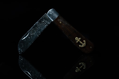 A folding pocket knife with a wooden handle and metallic blade is depicted. The handle features a brass anchor emblem and the blade has visible water droplets. The knife stands slightly open, reflecting on the dark surface beneath.