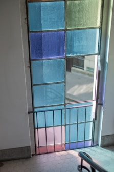 A door constructed with colorful stained glass panels in shades of blue, green, and purple. The door features a black metal frame and a gate at the bottom. Sunlight casts shadows on tiled flooring inside a building, creating a serene and artistic atmosphere.