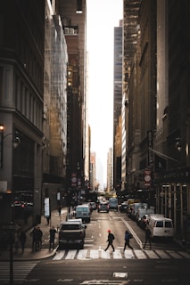 Tall skyscrapers line a bustling city street, with a mix of pedestrians crossing the road and vehicles driving through. The street is illuminated by the golden hour light, casting a warm glow on the buildings and creating long shadows. The atmosphere is dynamic and urban, with a sense of activity and movement.