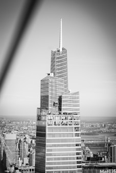 A modern skyscraper with a sleek, geometric design and a spire at the top. The building features glass panels that reflect the city skyline, which stretches into the background. The image captures a clear view of the urban landscape in black and white.