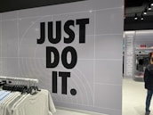 The image features a retail store interior with a prominent slogan 'JUST DO IT' in large black letters on a tiled wall. There is a clothing rack in the foreground with various T-shirts displayed on hangers. The wall has a grid pattern with subtle circular lines. To the right, a sign indicates fitting rooms, and a person is partially visible. The store is well-lit with modern fixtures.