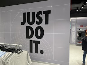 The image features a retail store interior with a prominent slogan 'JUST DO IT' in large black letters on a tiled wall. There is a clothing rack in the foreground with various T-shirts displayed on hangers. The wall has a grid pattern with subtle circular lines. To the right, a sign indicates fitting rooms, and a person is partially visible. The store is well-lit with modern fixtures.