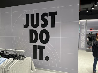 The image features a retail store interior with a prominent slogan 'JUST DO IT' in large black letters on a tiled wall. There is a clothing rack in the foreground with various T-shirts displayed on hangers. The wall has a grid pattern with subtle circular lines. To the right, a sign indicates fitting rooms, and a person is partially visible. The store is well-lit with modern fixtures.