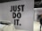 The image features a retail store interior with a prominent slogan 'JUST DO IT' in large black letters on a tiled wall. There is a clothing rack in the foreground with various T-shirts displayed on hangers. The wall has a grid pattern with subtle circular lines. To the right, a sign indicates fitting rooms, and a person is partially visible. The store is well-lit with modern fixtures.