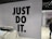 The image features a retail store interior with a prominent slogan 'JUST DO IT' in large black letters on a tiled wall. There is a clothing rack in the foreground with various T-shirts displayed on hangers. The wall has a grid pattern with subtle circular lines. To the right, a sign indicates fitting rooms, and a person is partially visible. The store is well-lit with modern fixtures.