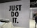 The image features a retail store interior with a prominent slogan 'JUST DO IT' in large black letters on a tiled wall. There is a clothing rack in the foreground with various T-shirts displayed on hangers. The wall has a grid pattern with subtle circular lines. To the right, a sign indicates fitting rooms, and a person is partially visible. The store is well-lit with modern fixtures.