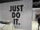 The image features a retail store interior with a prominent slogan 'JUST DO IT' in large black letters on a tiled wall. There is a clothing rack in the foreground with various T-shirts displayed on hangers. The wall has a grid pattern with subtle circular lines. To the right, a sign indicates fitting rooms, and a person is partially visible. The store is well-lit with modern fixtures.