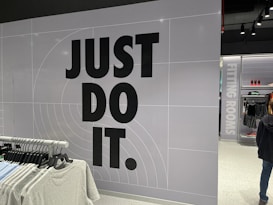 The image features a retail store interior with a prominent slogan 'JUST DO IT' in large black letters on a tiled wall. There is a clothing rack in the foreground with various T-shirts displayed on hangers. The wall has a grid pattern with subtle circular lines. To the right, a sign indicates fitting rooms, and a person is partially visible. The store is well-lit with modern fixtures.